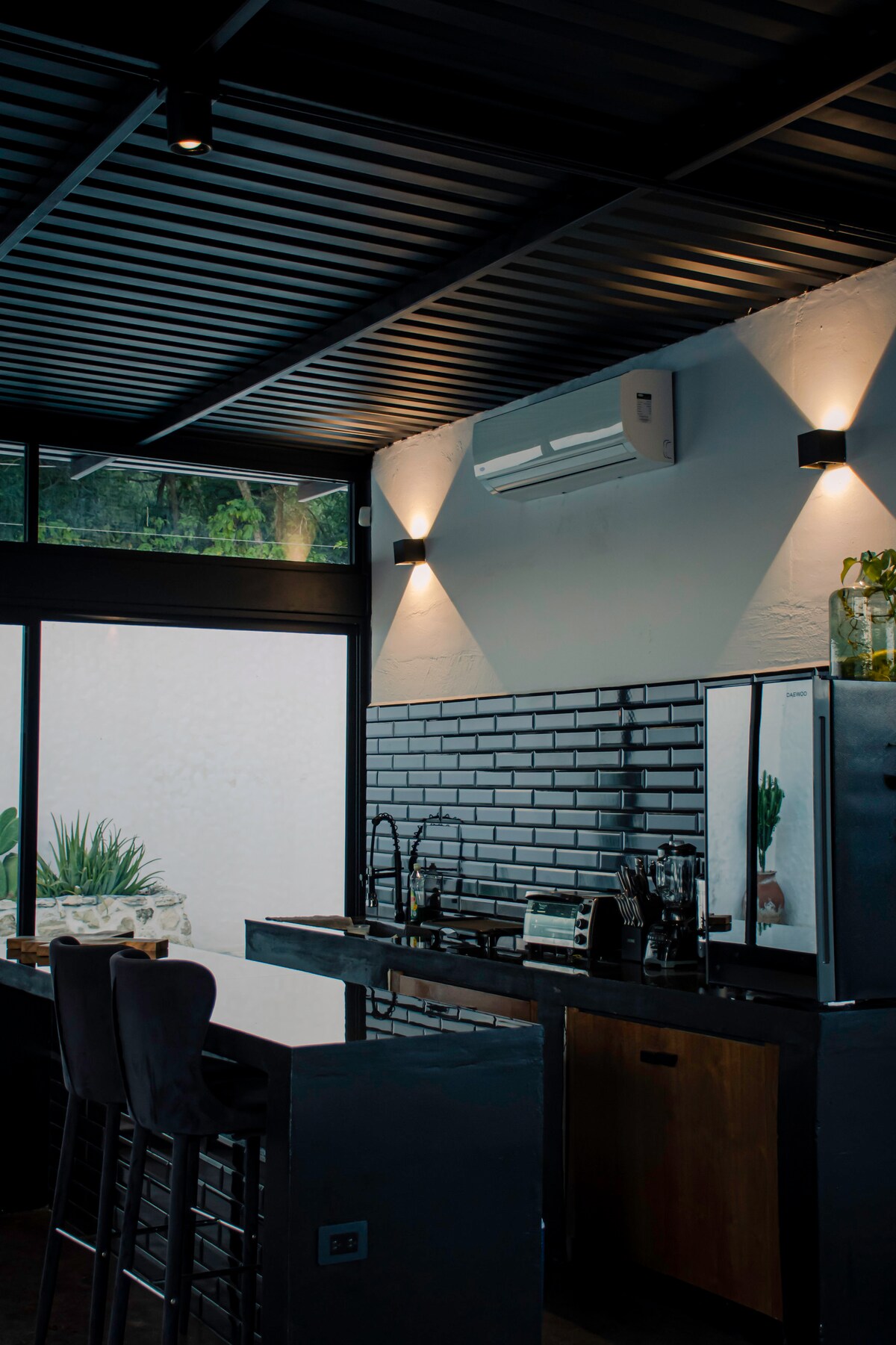 A modern kitchen space is showcased with sleek cabinetry and a dark countertop. Two high stools are positioned at the island. Large windows allow natural light to fill the area, while potted plants add a touch of greenery, enhancing the kitchen's fresh ambiance.