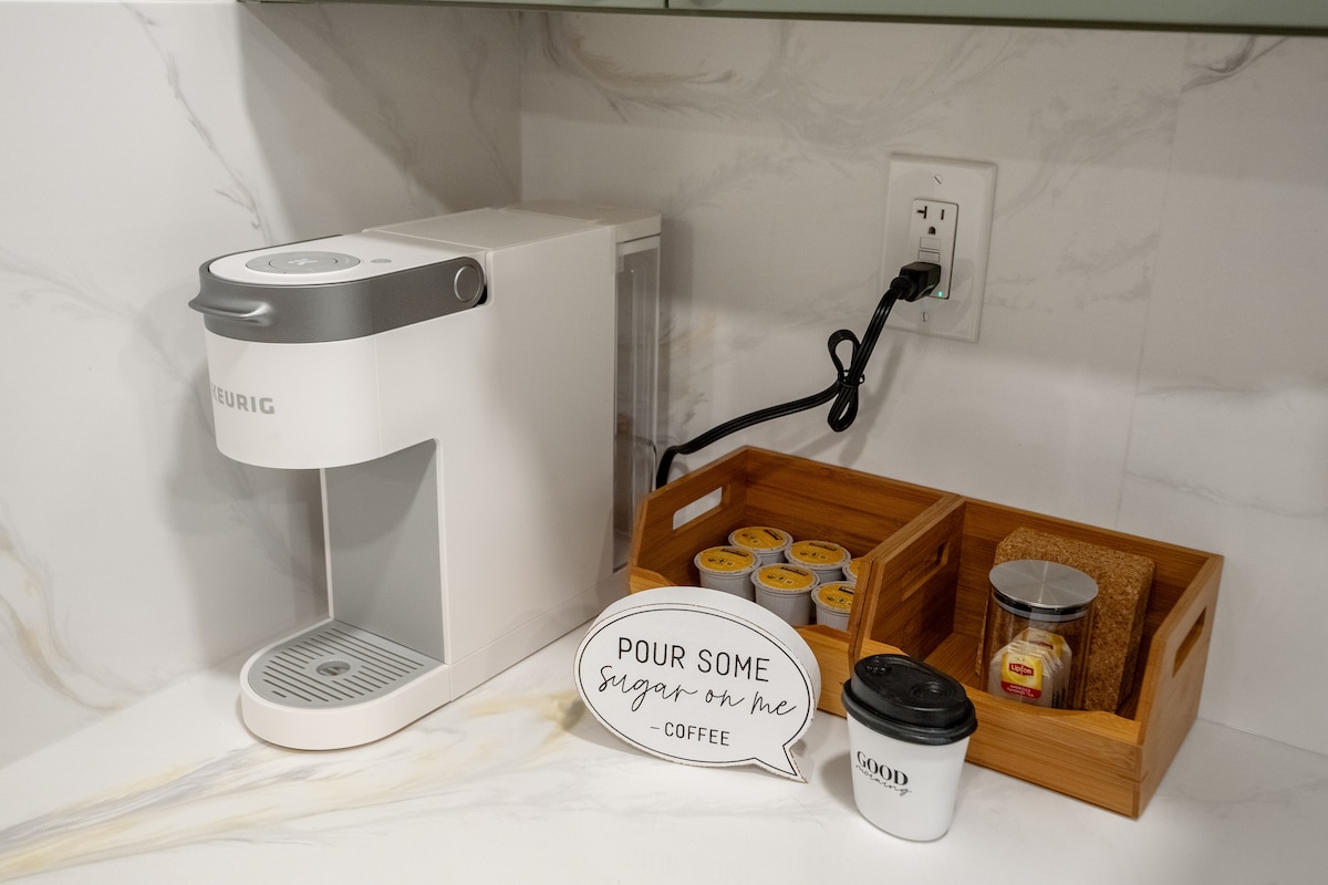 A coffee station is equipped with a Keurig machine, surrounded by wooden containers holding coffee pods and a sugar packet. A small cup with a lid is placed nearby, along with a charming sign displaying the phrase 'Pour some sugar on me - coffee.'