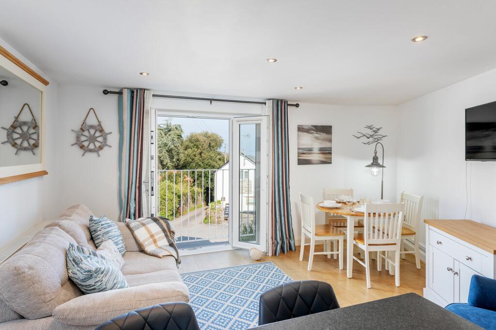 The open plan living and dining area features a light-colored sofa adorned with cushions. A wooden dining table with white chairs is positioned nearby. Juliet-style doors lead out to a breezy balcony area, allowing natural light to fill the space.