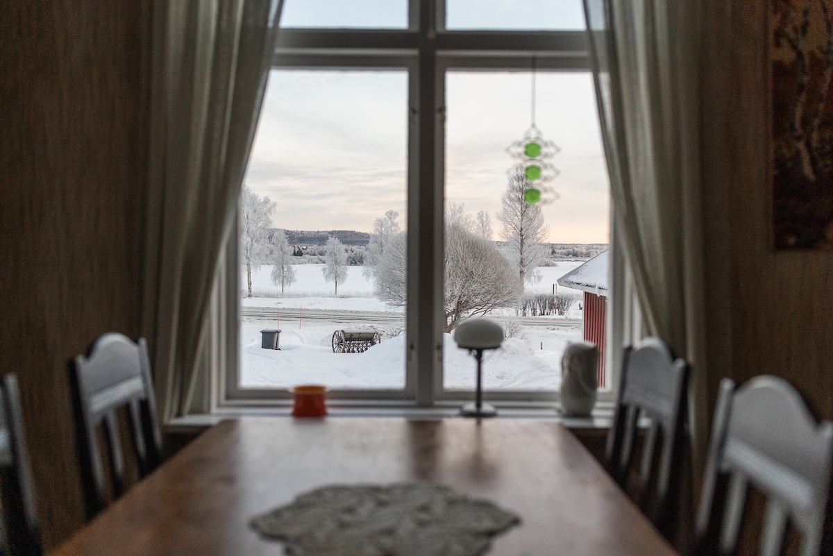A dining table is positioned in front of a large window offering a view of a snow-covered landscape. Frosted trees line the foreground, and a subtle glow of soft light is visible beyond. A decorative pendant light hangs above the table, adding a touch of warmth.