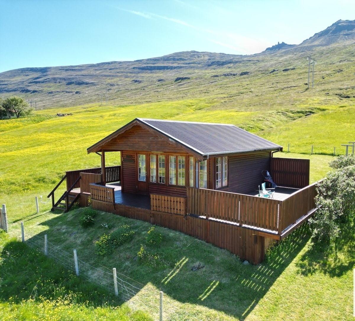 A cozy wooden cabin is set against a backdrop of rolling green hills. Its exterior features a wraparound deck with railings, while large windows offer glimpses of the interior. The sunny landscape enhances the inviting presence of the cabin.
