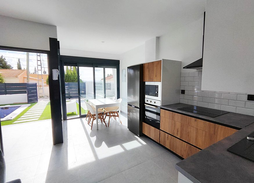 A modern kitchen space includes sleek cabinetry in wood and gray tones. Sunlight filters through large glass doors, illuminating the dining area with a white table and wooden chairs. An outdoor pool area and lush greenery can be seen through the doors, enhancing the serene ambiance.