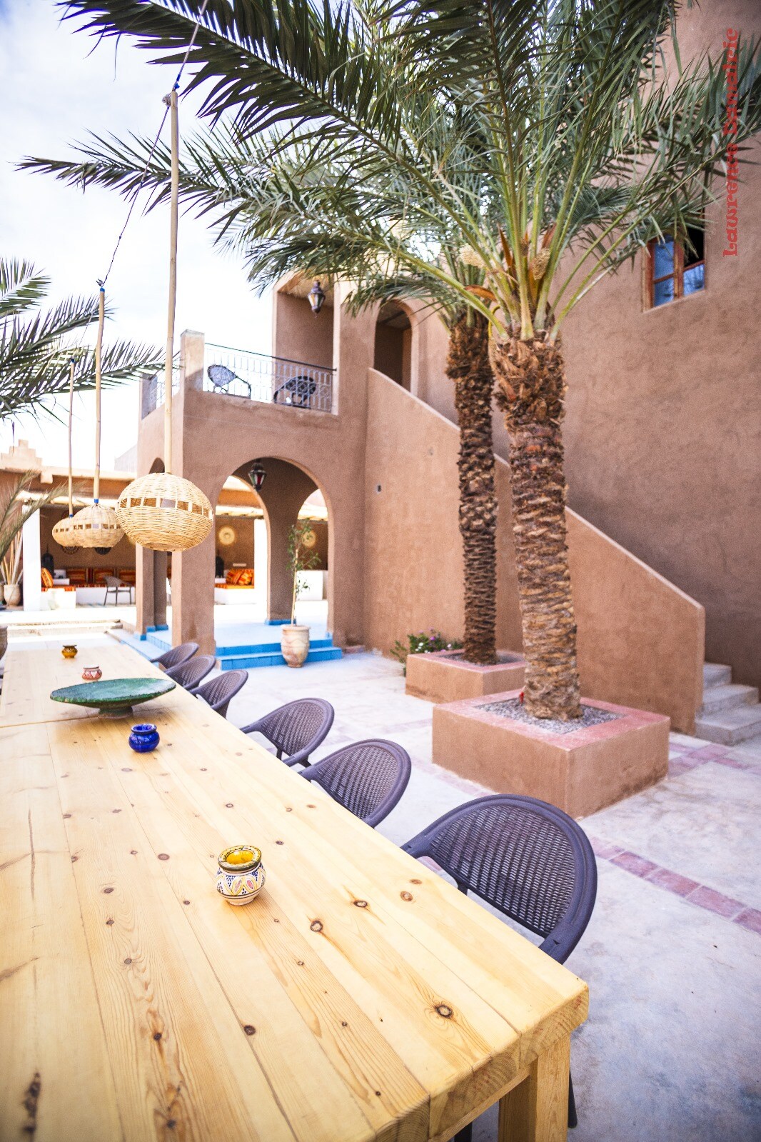 A spacious outdoor dining area features a long wooden table surrounded by dark chairs. Palm trees provide shade, and a charming staircase and archways are visible in the background. The earthy tones of the building complement the vibrant greenery.