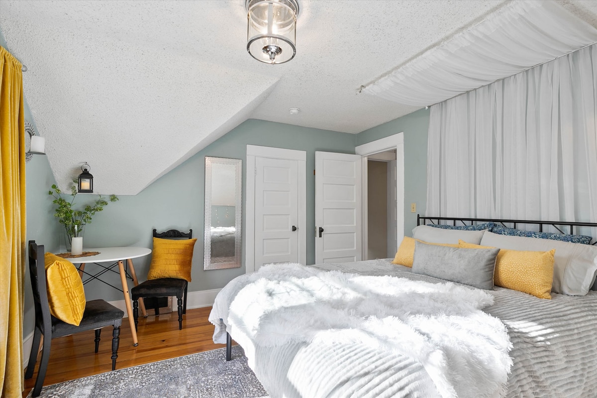 A bright bedroom featuring a spacious bed adorned with textured blankets and accent pillows. A chair and small table are positioned nearby, along with a potted plant. Natural light filters in through light curtains, and a mirrored surface adds depth to the space.