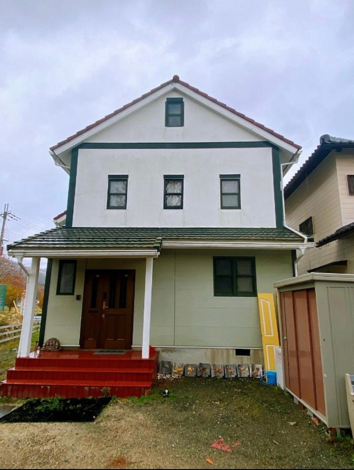 A two-story house features a traditional design with a white exterior and dark green trim. The entrance is highlighted by a brown door, and a small set of stairs leads up to it. Nearby, a storage shed adds functionality to the outdoor space.