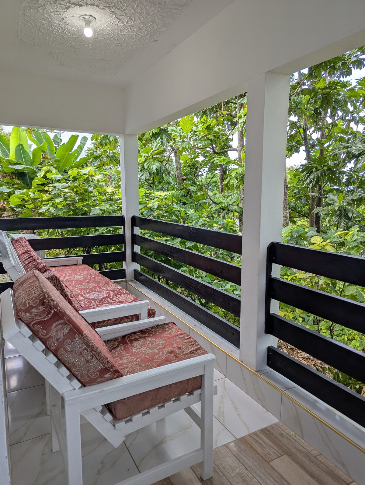 A covered balcony provides a serene view of lush greenery, featuring two white wooden chairs with red patterned cushions. The space is surrounded by plants, creating a peaceful and natural atmosphere.