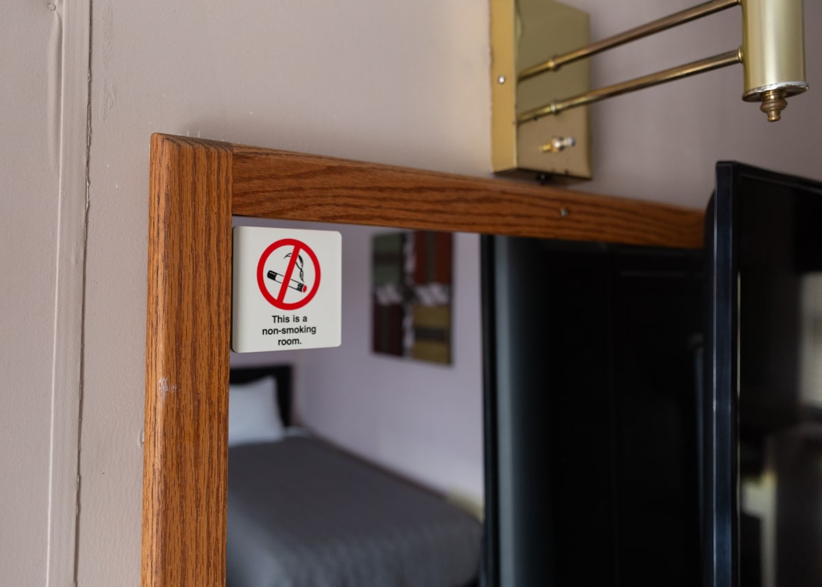 A mirror is framed in wood, with a notice indicating the room is non-smoking. In the background, a spacious bed is visible, along with a hint of colorful artwork on the wall, adding to the room's character.