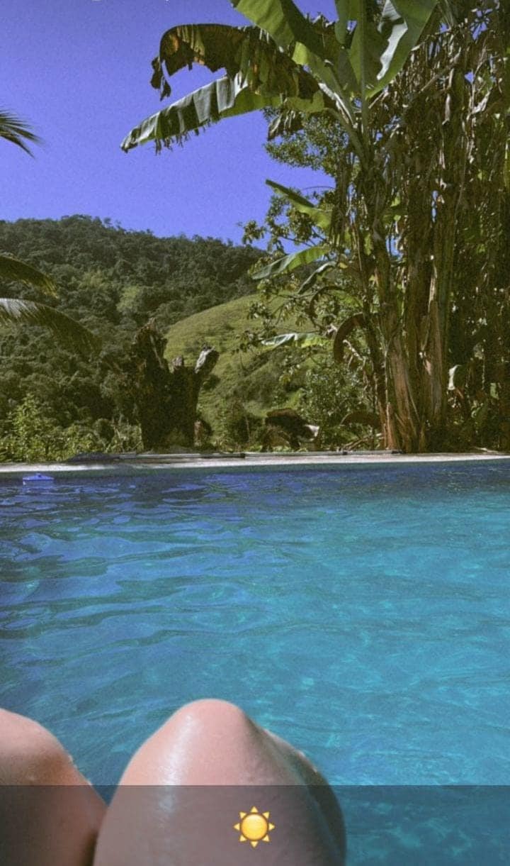 A clear swimming pool glistens in the sunlight, surrounded by lush greenery and tropical plants. The serene view of a hillside is visible in the background, providing a sense of tranquility and connection to nature.