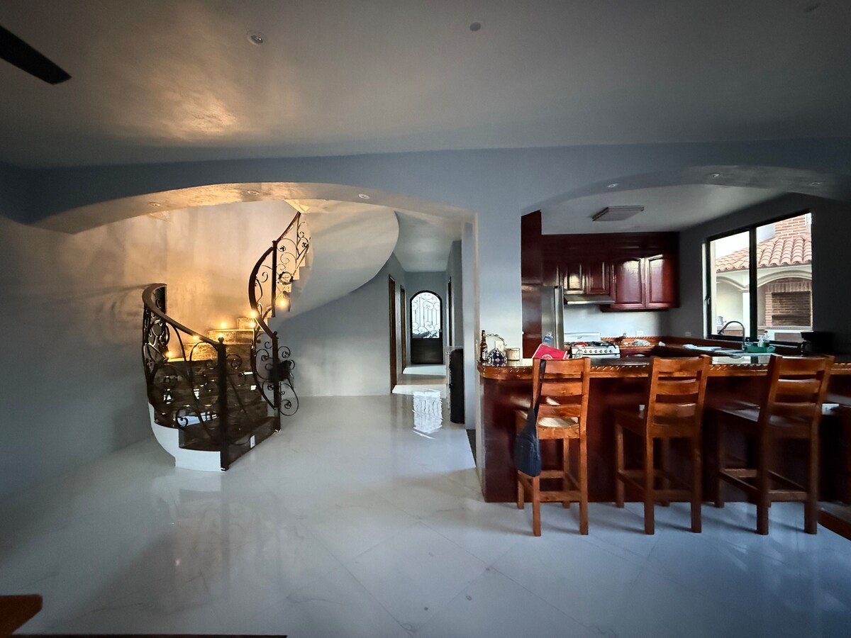 A modern kitchen with dark wood cabinetry and a spacious breakfast bar is visible. Natural light fills the area through large windows. Curved stairs with decorative iron railings lead to an upper level, enhancing the open feel of the space.