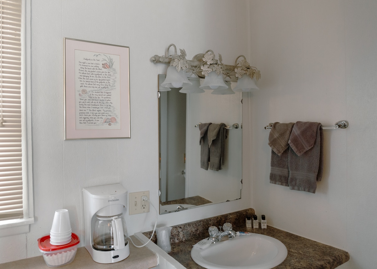 The bathroom features a clean sink area with a white basin. A large mirror is mounted above the sink, flanked by three light fixtures. A coffee maker and disposable cups are placed on the countertop, alongside toiletries. Fresh towels are neatly arranged on a rack.