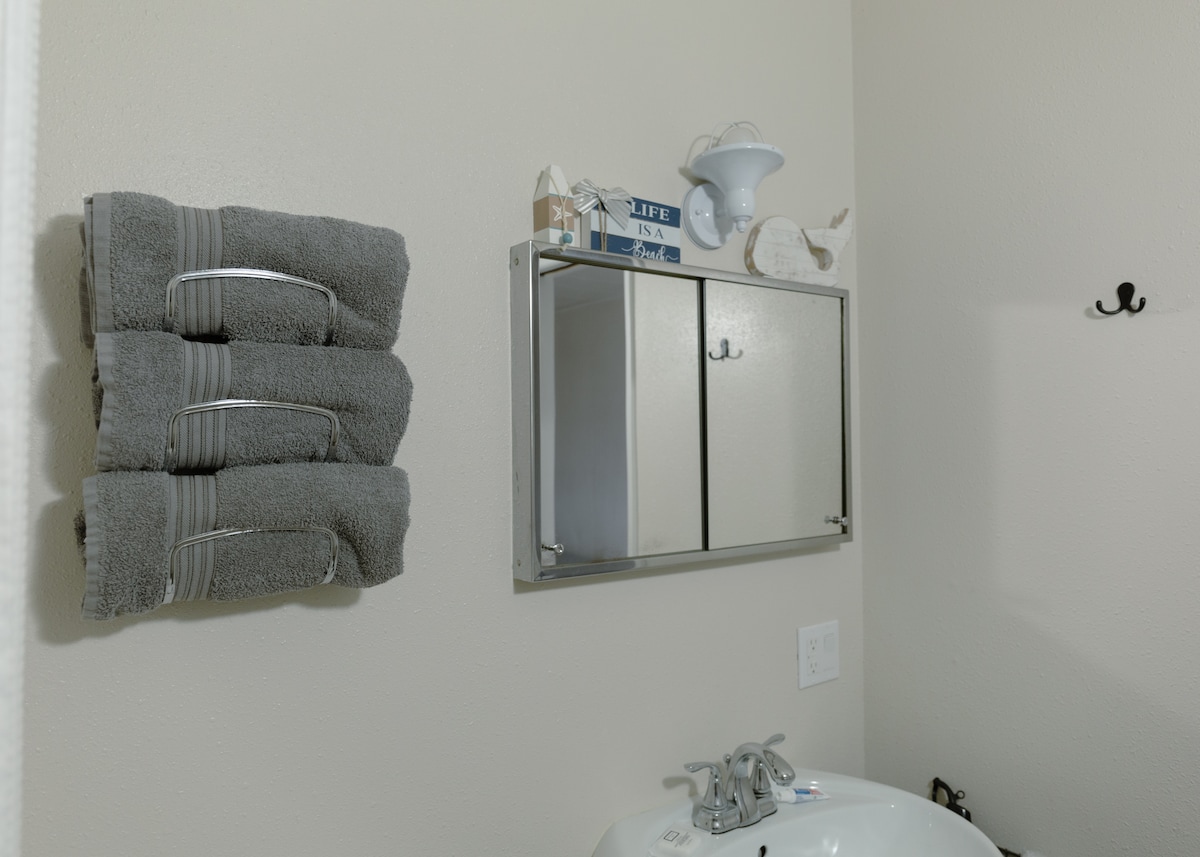 A clean and functional bathroom features neatly stacked gray towels on a rack. A mirrored cabinet is mounted above the sink, providing storage space. A subtle wall hook is visible, complementing the overall organization of the area.