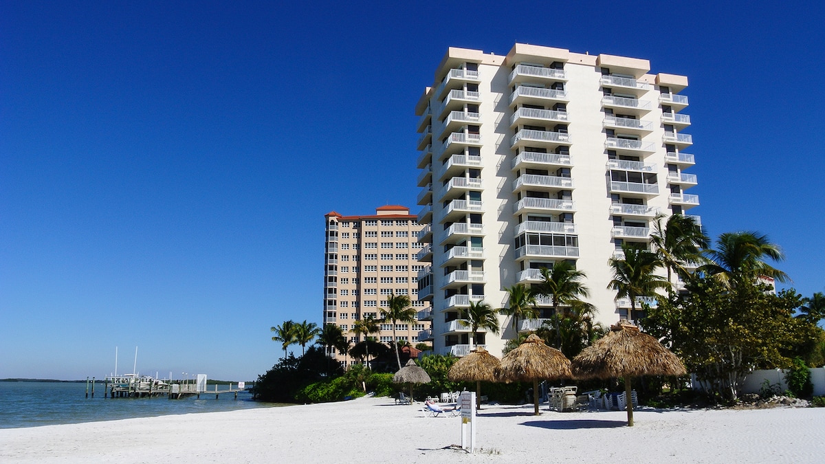 A beachfront view of a multi-story condo building, complemented by palm trees and straw umbrellas on the sandy shore. Clear blue skies and the adjacent pier extend towards the water, enhancing the coastal atmosphere.