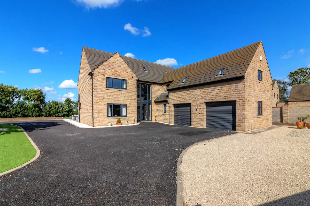 The exterior of a large stone house features a mix of natural stone and modern design elements. A spacious driveway with ample parking leads to the double garage. Lush greenery surrounds the property, and large windows reflect the clear blue sky.