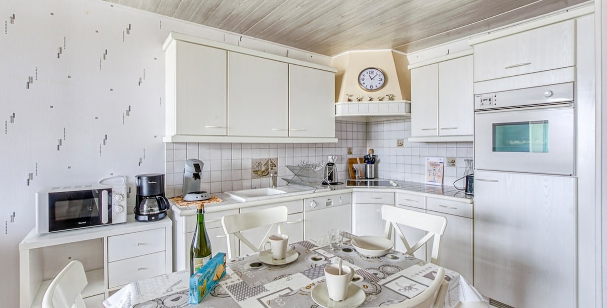 A bright kitchen area includes modern white cabinetry and tiled countertops. A dining table with white chairs is set with dishes and beverages. Appliances such as a microwave, coffee maker, and oven are visible, along with a wall clock and various cooking essentials.