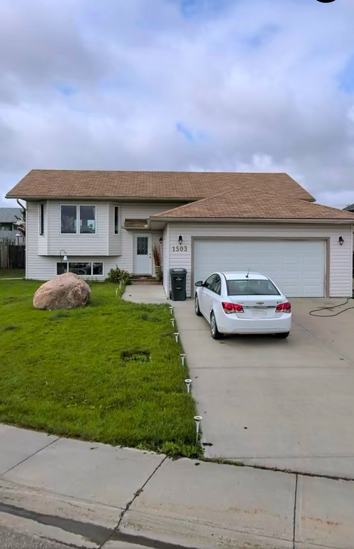 The exterior of a two-story house is depicted, featuring a beige facade and a brown shingled roof. A large boulder is placed on the front lawn, and a driveway accommodates a white car parked beside a garage. Lush green grass surrounds the home.