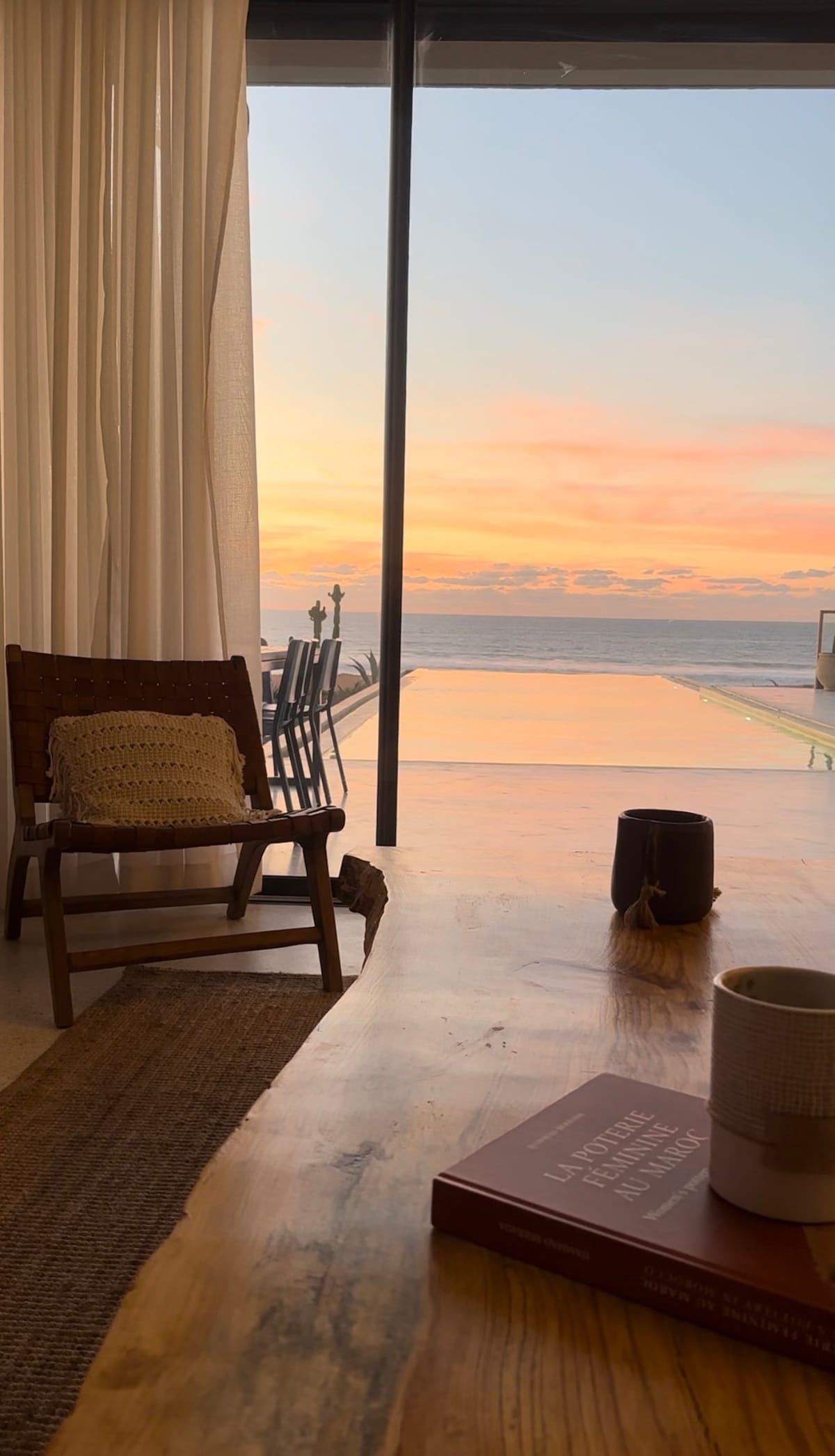 A spacious living area is visible, featuring a wooden table and a cozy chair adorned with a knitted cushion. Large glass doors open to reveal a sunset over the sea, with the reflection on the water creating a serene ambiance. Soft curtains drape alongside the doorway.