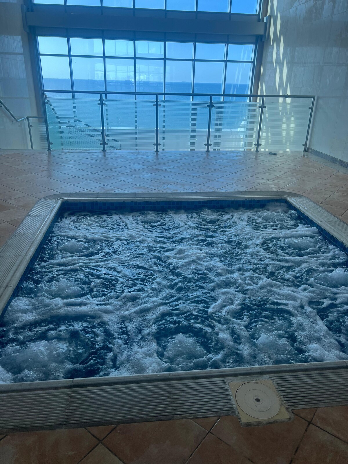 A spacious hot tub is situated indoors, with bubbling water creating an inviting scene. Large glass doors nearby allow natural light to flood the area, while views of the sea are visible beyond a glass barrier. The tiled floor complements the modern aesthetic.