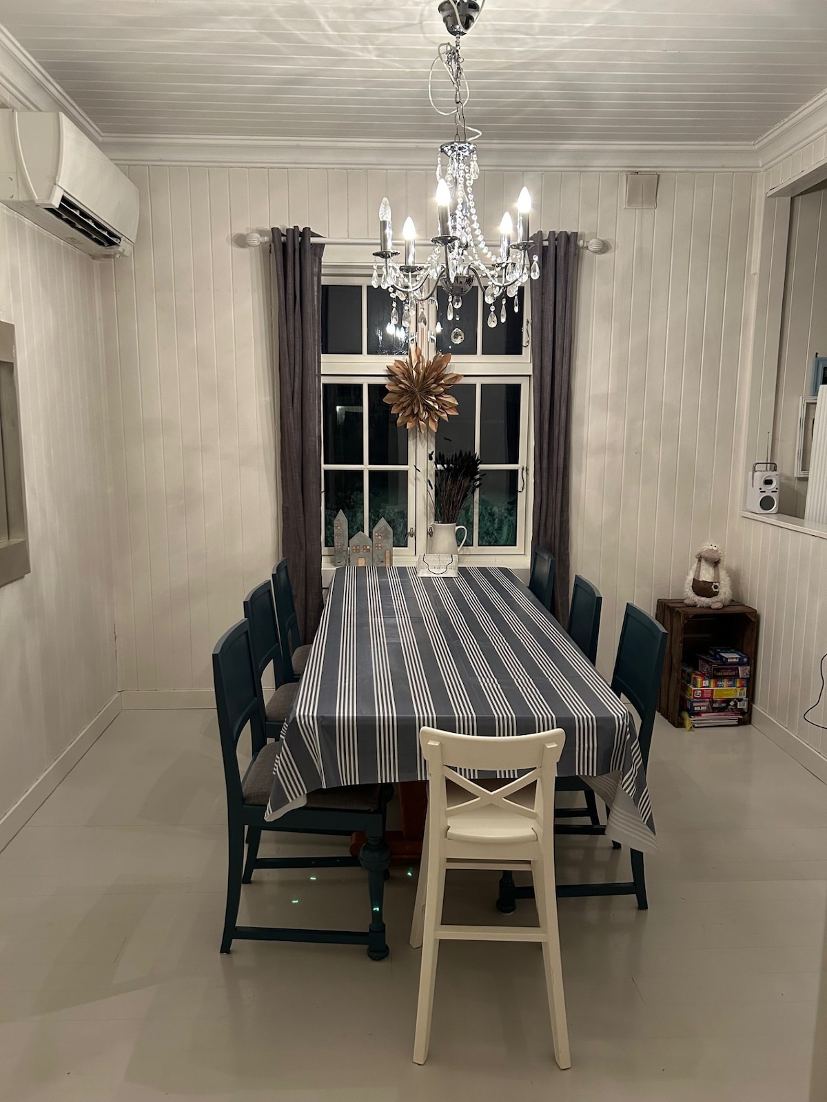 A dining area features a long table surrounded by colorful chairs. A chandelier hangs overhead, providing illumination. Light neutral walls and a large window create an open feel, with curtains offering some privacy. A small side table displays games and decorative items.