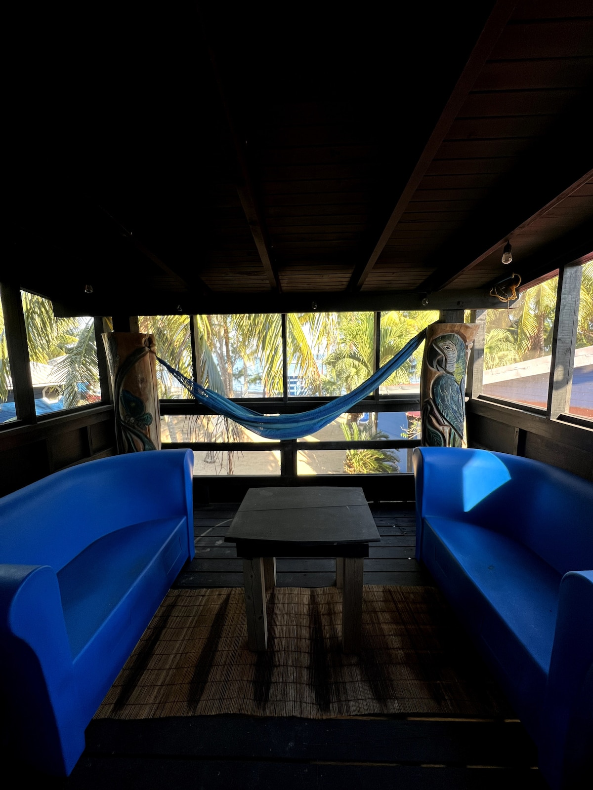 An indoor seating area features two blue couches facing a central table. Colorful decor on the walls adds character while large windows provide views of the palm-fringed exterior. A hammock is positioned between the windows, inviting relaxation in a bright space filled with natural light.