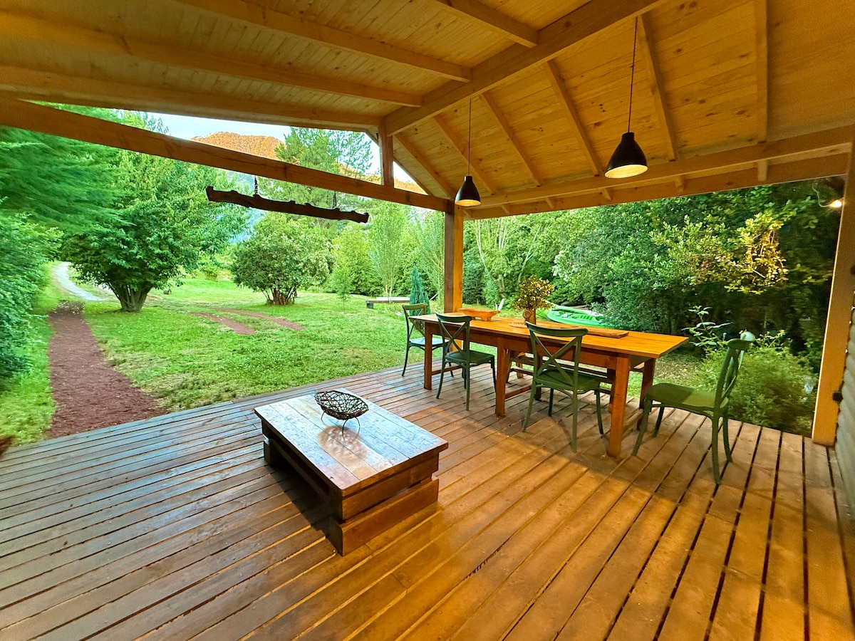 An inviting outdoor dining area features a wooden table surrounded by green chairs. A rustic wooden coffee table sits nearby, complemented by shaded lighting fixtures overhead. Lush greenery and a gravel pathway create a peaceful atmosphere, enhancing the connection to nature.