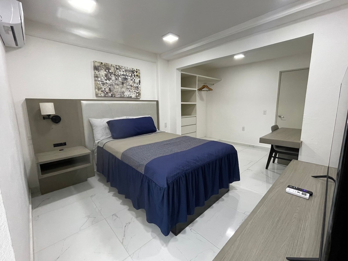 A comfortable sleeping area features a spacious bed with a dark blue bedspread. A light-colored accent wall is complemented by a decorative panel. A small desk and a television are visible, along with a closet that provides ample storage space.