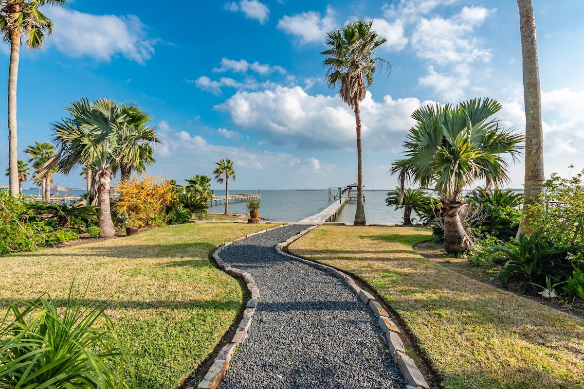 A winding stone pathway leads through manicured grassy areas and palm trees towards the waterfront. The distant fishing pier is visible under a bright sky dotted with clouds, creating a serene outdoor setting.