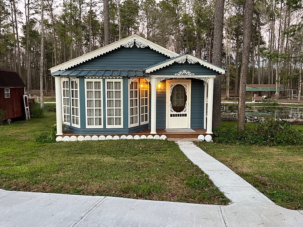 The exterior of a charming blue cottage is presented, featuring decorative white trim and large windows that invite natural light. A welcoming porch is adorned with intricate details, and a pathway leads to the entrance, surrounded by lush greenery and tall trees.