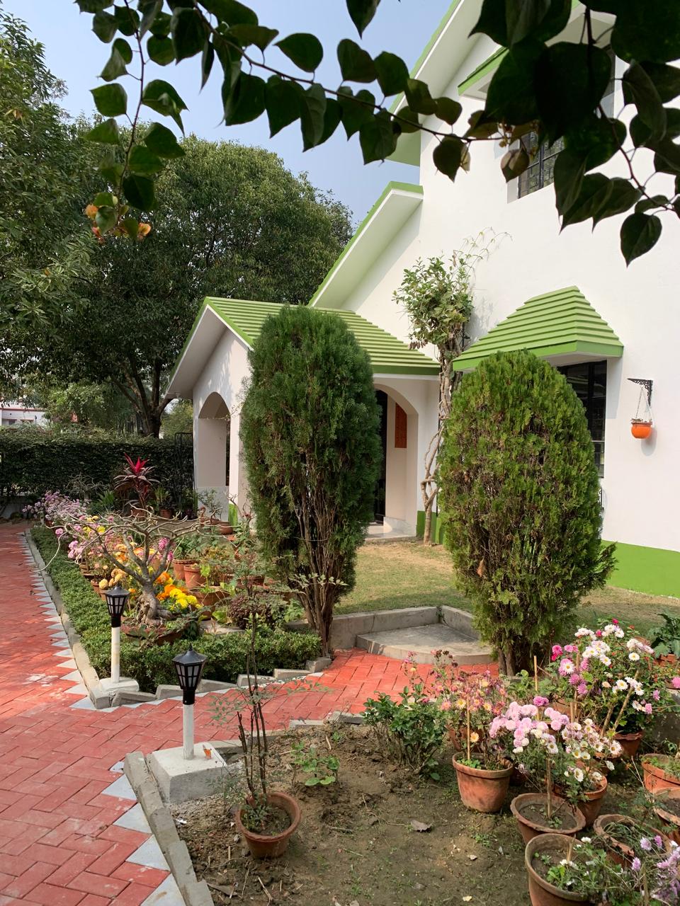 A charming pathway lined with flowering plants leads to the entrance of the villa. Flanking the pathway are tall green shrubs and decorative lights, creating an inviting entryway. The house features a sloped green roof and an arched entry, set against a backdrop of lush greenery.