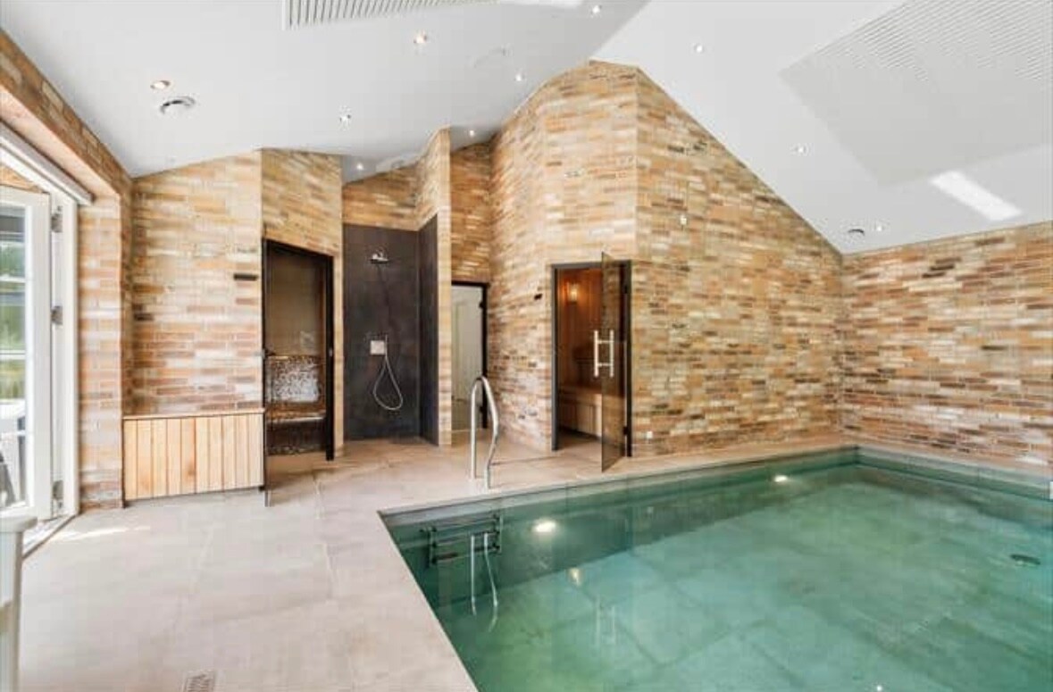 An indoor pool area features a saltwater pool with steps leading in. The surrounding walls are adorned with natural stone tiles, creating a warm atmosphere. Glass doors offer access to outdoor spaces, and areas for relaxation are present, including shower facilities and a steam room.