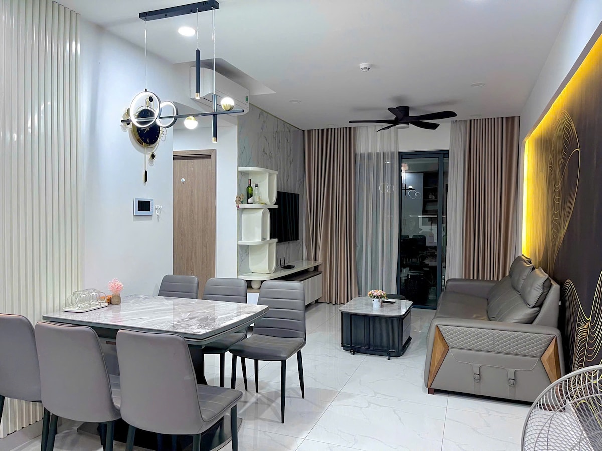 A modern living space is presented with a sleek dining table surrounded by six chairs. A comfortable seating area is located adjacent, featuring a grey sofa and a coffee table. Soft lighting and decorative wall accents enhance the room's ambiance, complemented by large windows dressed with soft curtains.
