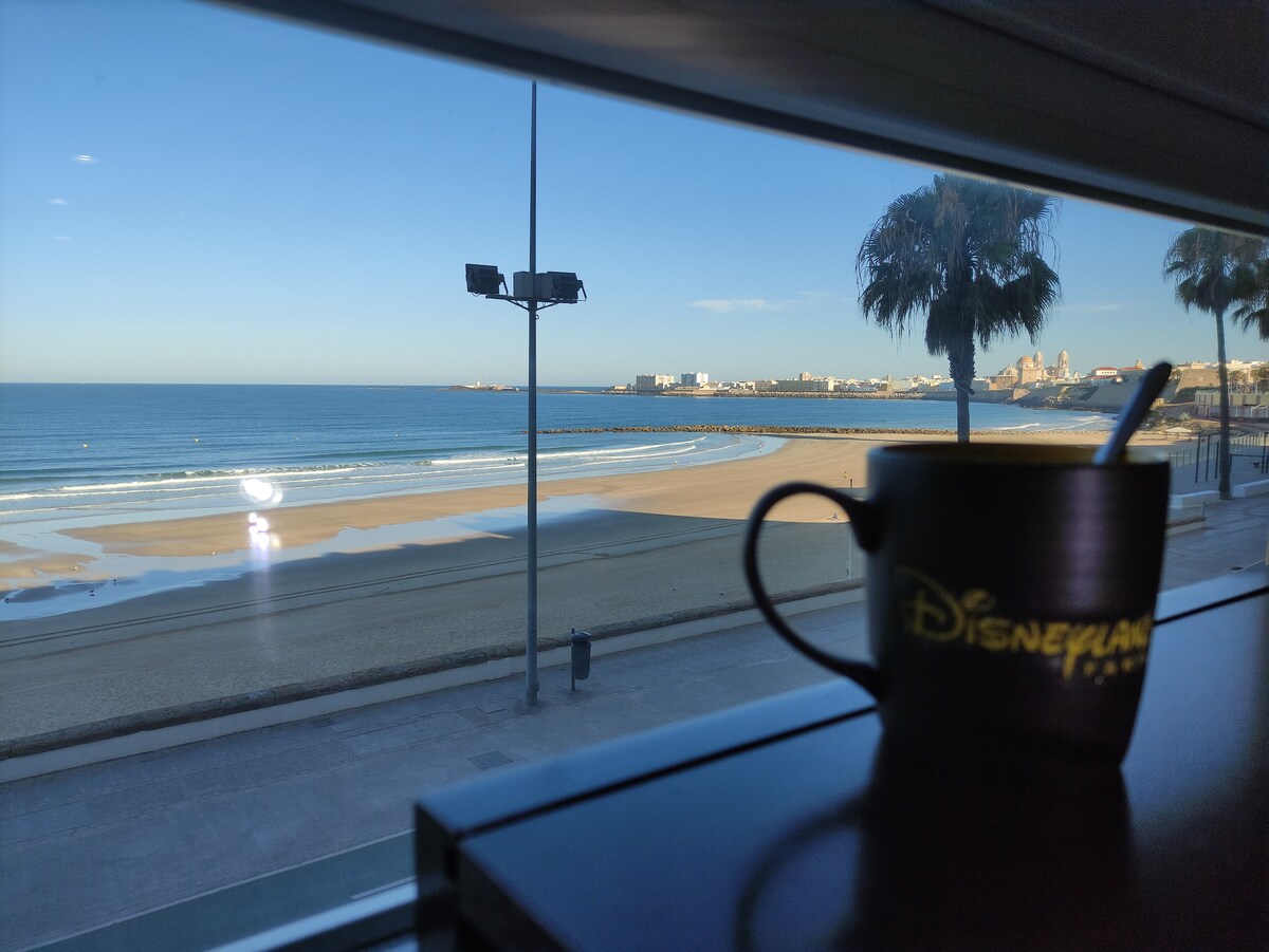 A coastal view is framed by a window, showcasing the sandy beach and gentle waves of Santa María del Mar. A mug, featuring a logo, sits on a ledge in the foreground, with palm trees swaying slightly in the breeze and a clear blue sky overhead.