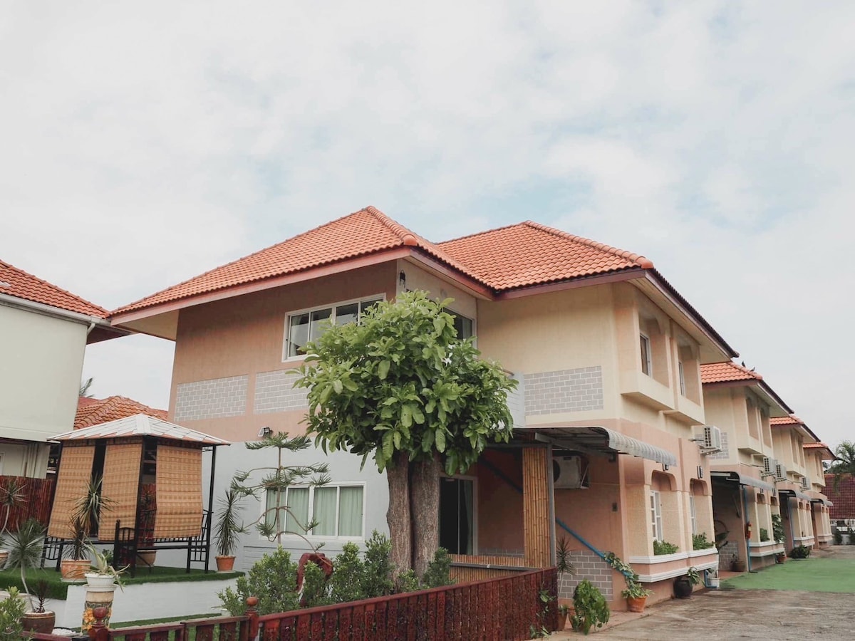 A two-story villa is featured with a distinctive red-tiled roof and light-colored exterior walls. The greenery includes a sizable tree in the front yard, while a shaded gazebo is visible on the left, providing a serene outdoor gathering space.
