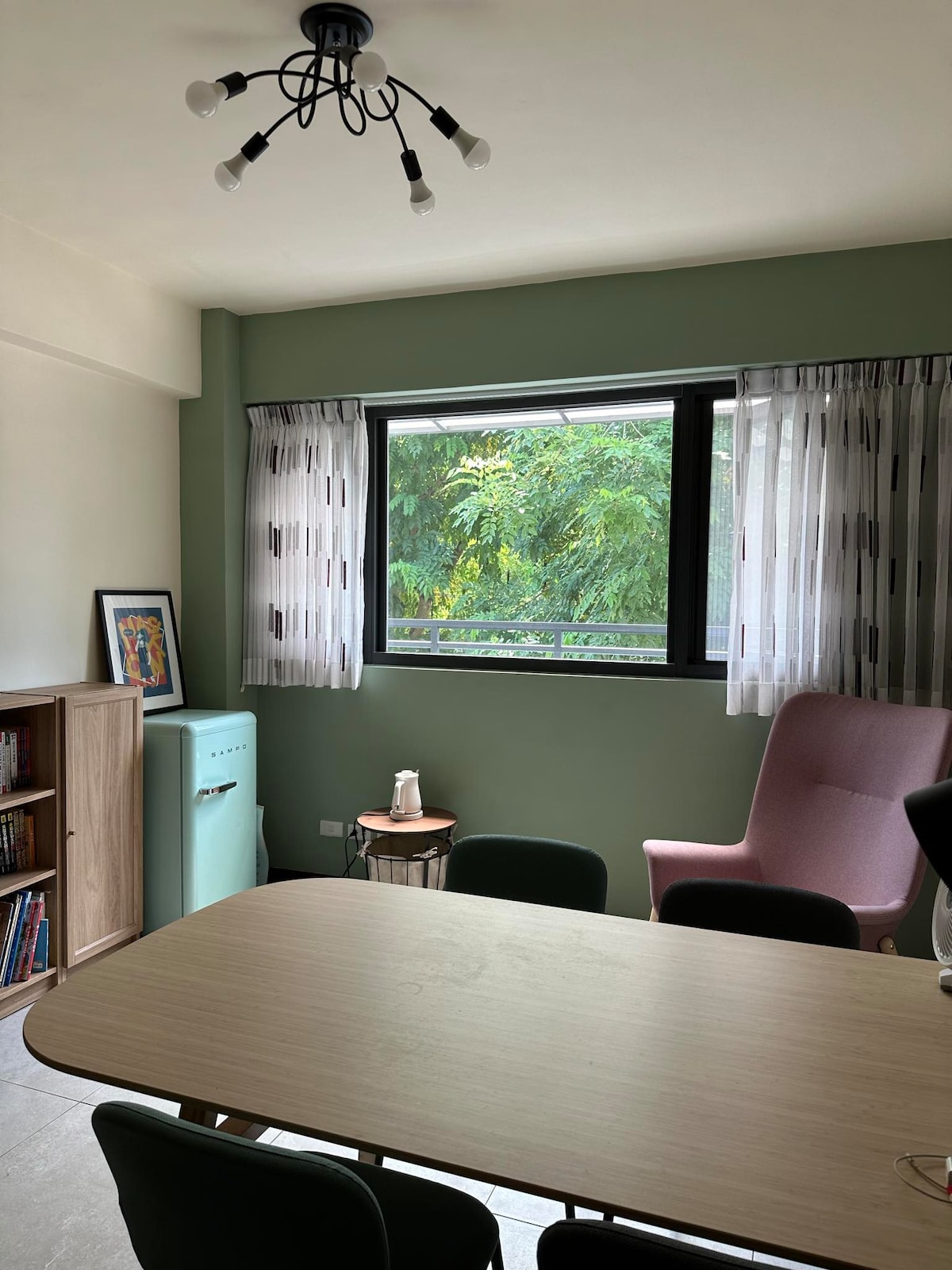 A comfortable dining area features a light wooden table surrounded by black chairs. A pink armchair sits beside a small side table, and a mint-green refrigerator stands against the wall. Natural light filters through a window dressed in sheer curtains, revealing greenery outside.