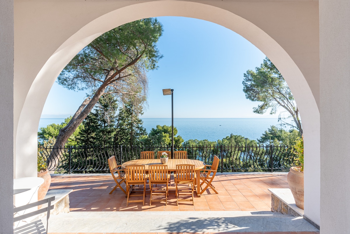 An outdoor terrace is seen under a large archway, featuring a wooden dining table with matching chairs. Lush green trees frame the panoramic sea view, highlighting the tranquil atmosphere of the surrounding landscape. Bright blue skies complement the serene setting.