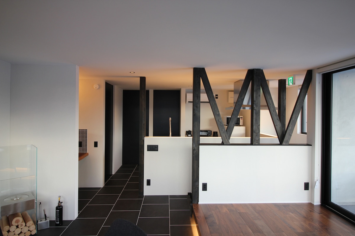 An open-plan living area is displayed with sleek black and wooden design elements. A modern kitchen is visible in the background. Large glass windows provide natural light and a clear view of the surroundings. Textured flooring adds depth to the space.
