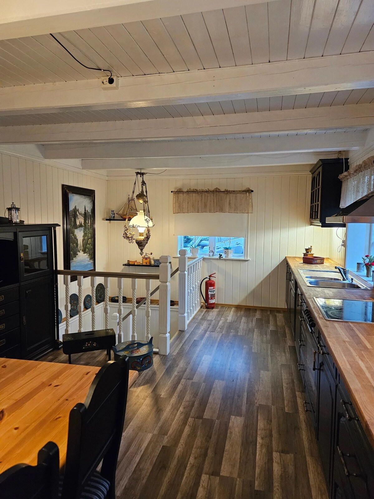 The kitchen features wooden countertops and black cabinetry, creating a warm and inviting space. Natural light enters through a window adorned with a simple curtain. A dining area with black chairs and a wooden table is visible, along with decorative artwork and a safety equipment display.
