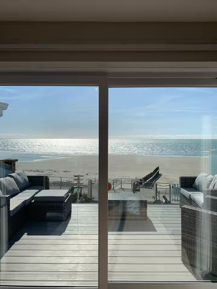 A view through glass doors captures sparkling water reflecting sunlight, extending towards a sandy beach. Comfortable outdoor seating is positioned on a wooden deck, inviting relaxation while enjoying the scenery. The distant beachfront is complemented by gentle waves and a clear sky.