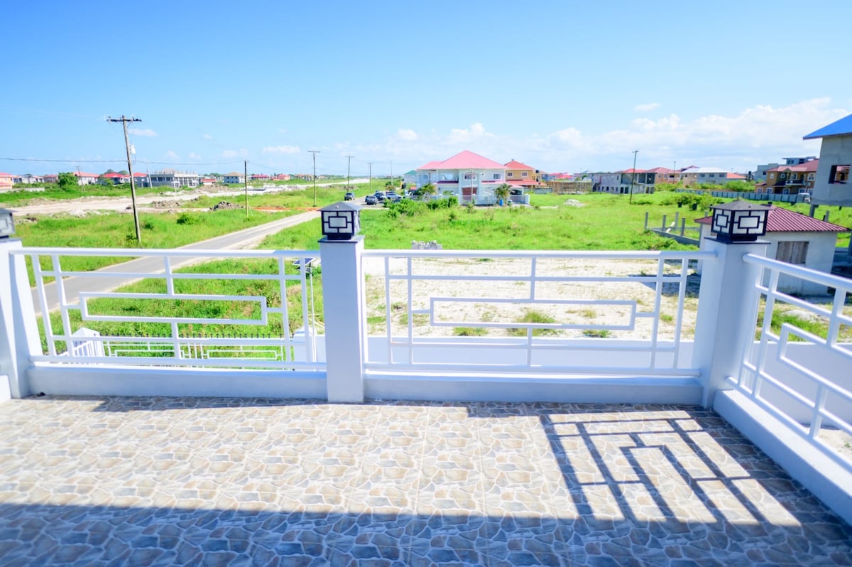 A clear view from a spacious patio is presented, featuring a decorative railing and a stone-patterned floor. Expansive green fields and distant buildings are visible across a quiet road under a bright blue sky, creating an open and airy atmosphere.
