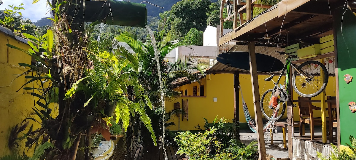 An outdoor area is surrounded by lush green plants, featuring a vibrant yellow wall. A hammock is suspended nearby, and a bicycle hangs from the wooden structure. Natural elements create a serene and private atmosphere within the space.