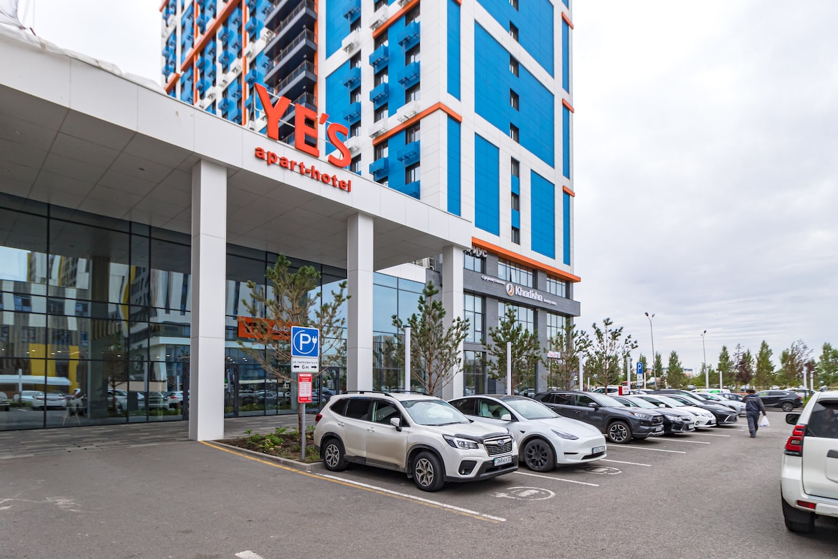 The exterior of the building features a modern facade with large glass windows and the 'YES apart-hotel' sign prominently displayed. A spacious parking area is visible, accommodating several vehicles, with greenery enhancing the entrance area.