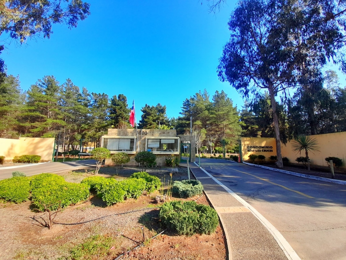 The entrance to Condominio Alto Las Cruces is shown, featuring a gated area with a notable Chilean flag. Well-maintained greenery surrounds the entrance, with tall trees in the background under a clear blue sky. A paved road leads into the area, indicating access to the complex.