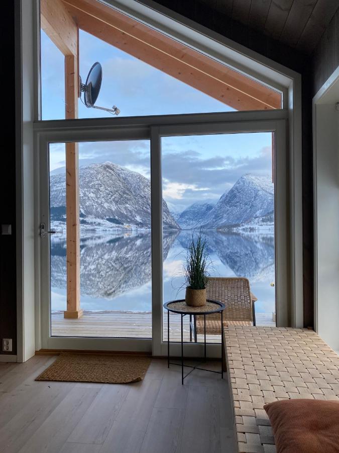 A large glass door offers stunning views of the snow-covered mountains reflecting in the calm water of Jølstravatnet. A small table with a potted plant is placed beside a woven chair. Natural light fills the interior space through the expansive window.