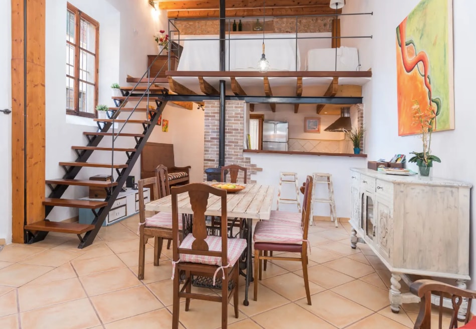 An open-plan living space features a dining table surrounded by wooden chairs. A wooden staircase leads to a loft area, showcasing high ceilings and a bright, airy atmosphere. Natural light enters through large windows, enhancing the rustic charm of the interior.