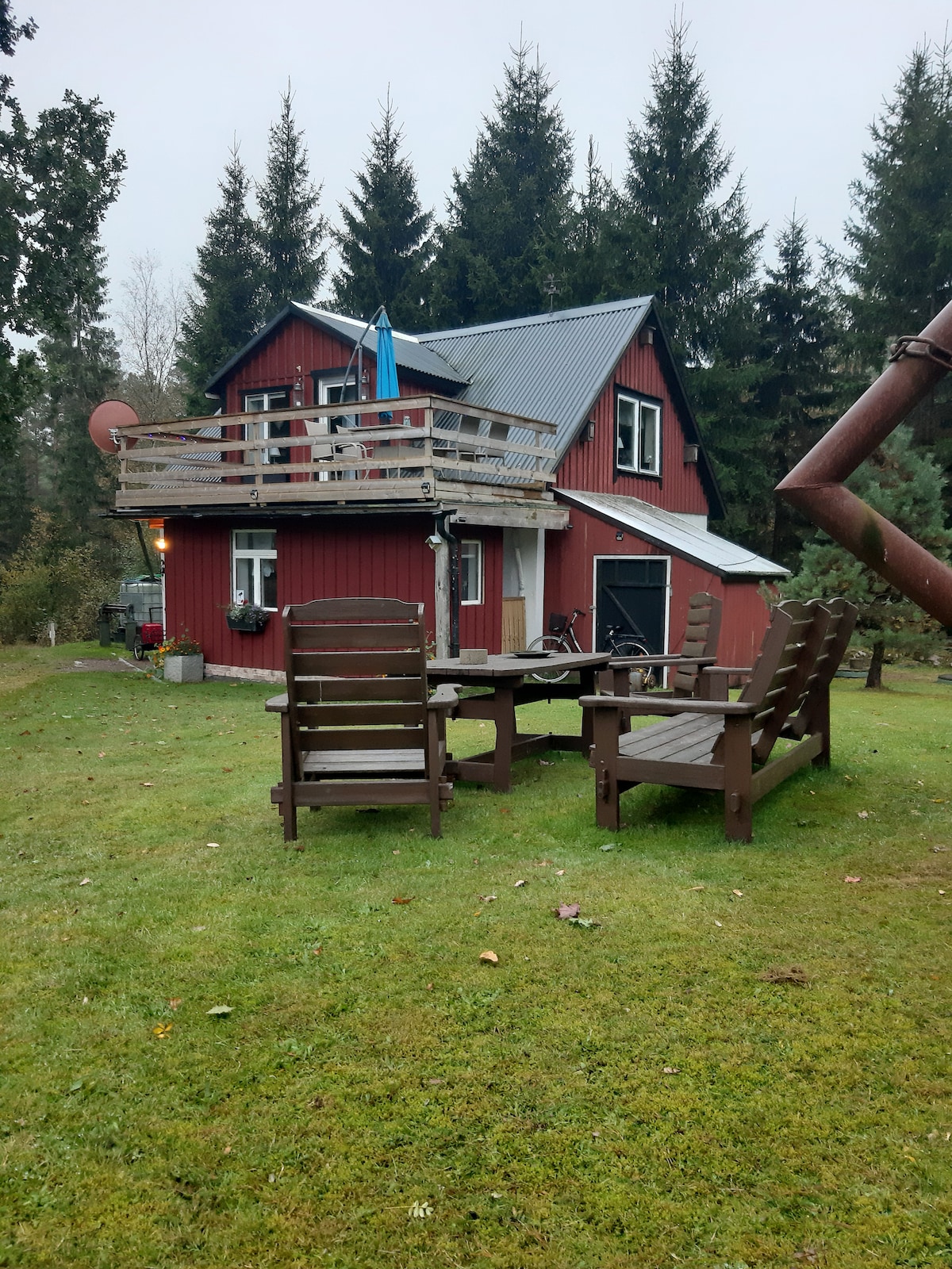 A quaint red cabin is surrounded by lush greenery. A cozy seating area made of wooden chairs stands on the lawn, ideal for relaxation. The cabin features a second-story balcony with a blue flag, and multiple windows allow natural light to fill the interior.
