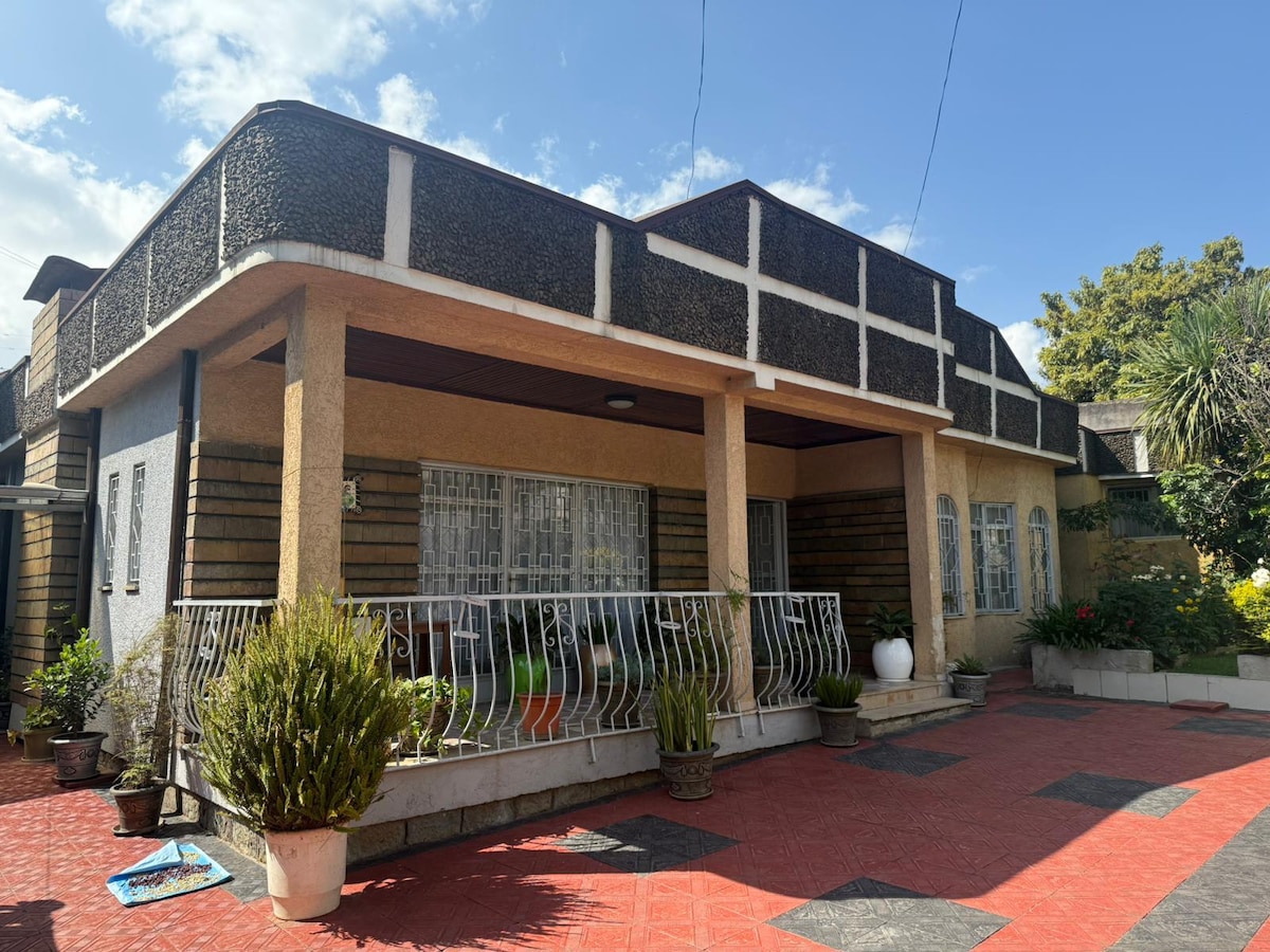 The exterior of the home features a spacious front porch adorned with decorative plants in pots. The building showcases a blend of textured walls and a wide balcony, complemented by large windows that allow for ample natural light. A red tiled pathway leads to the entrance.