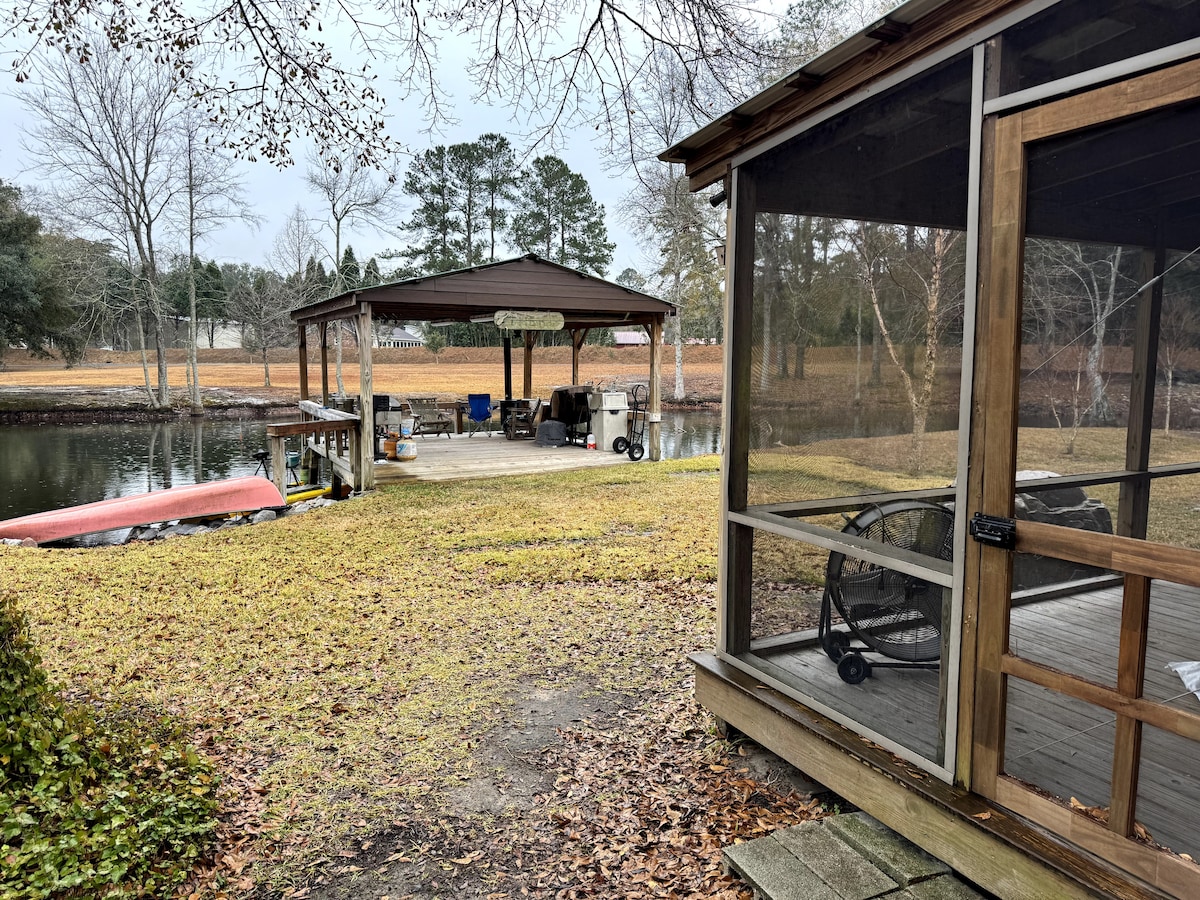 A view of a private dock set alongside a tranquil pond, with shaded seating and a workspace area visible under a protective roof. Surrounding greenery and bare trees indicate a serene outdoor environment, while the gentle slope of the lawn leads to the water's edge.
