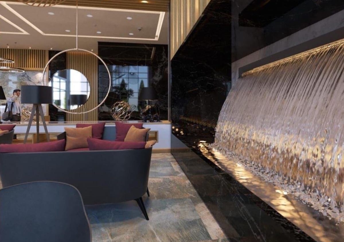 A modern lobby features a sleek water wall cascading beside a cozy seating area with dark chairs and colorful cushions. Accent lighting highlights the interplay of textures, while a reception desk is visible in the background, enhancing the inviting ambiance.