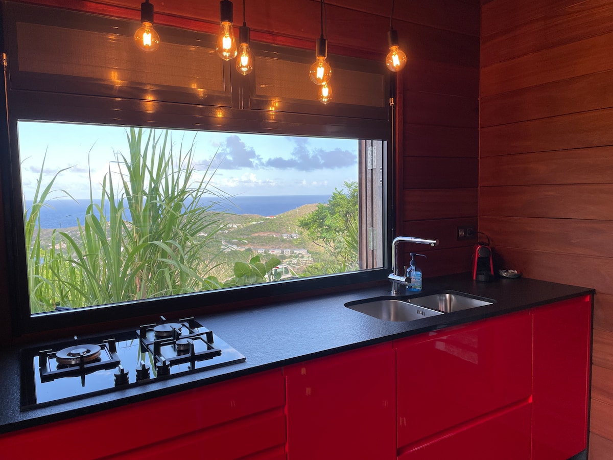 A modern kitchen features a sleek red cabinetry and a black countertop. A large window frames a stunning view of the surrounding landscape, with greenery visible outside. Pendant lights hang above the cooking area, which includes a gas stove and a stainless-steel sink.
