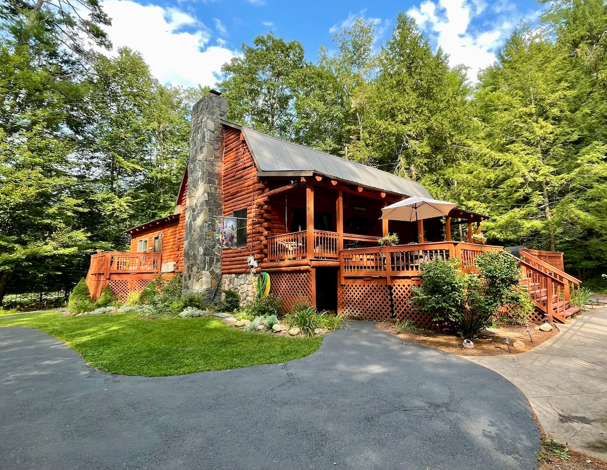 A rustic cabin is framed by lush greenery, featuring a prominent stone chimney and a spacious porch with railings. The exterior combines wooden textures with a roof under a clear blue sky, offering a sense of tranquility and connection with nature.