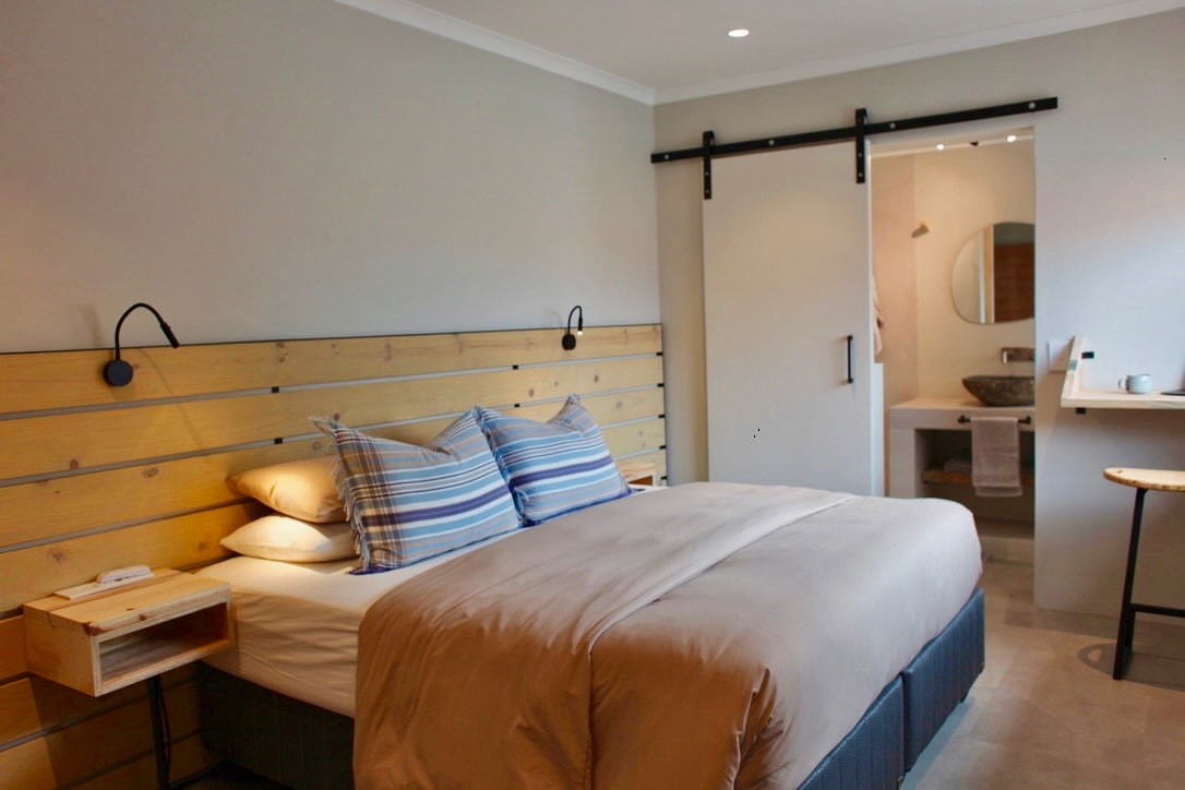 A spacious sleeping area features a comfortable bed with layered bedding and striped pillows. Bedside tables are positioned on either side, and a sliding door leads to a private bathroom. Natural light fills the room, accentuating the neutral color palette and wood accents.